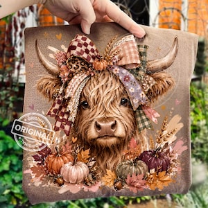 May include: A brown t-shirt featuring a graphic of a Highland cow with a large, multi-patterned bow between its horns. The cow is surrounded by autumn leaves, wheat stalks, and pumpkins in shades of orange, green, and purple. The design includes small hearts.