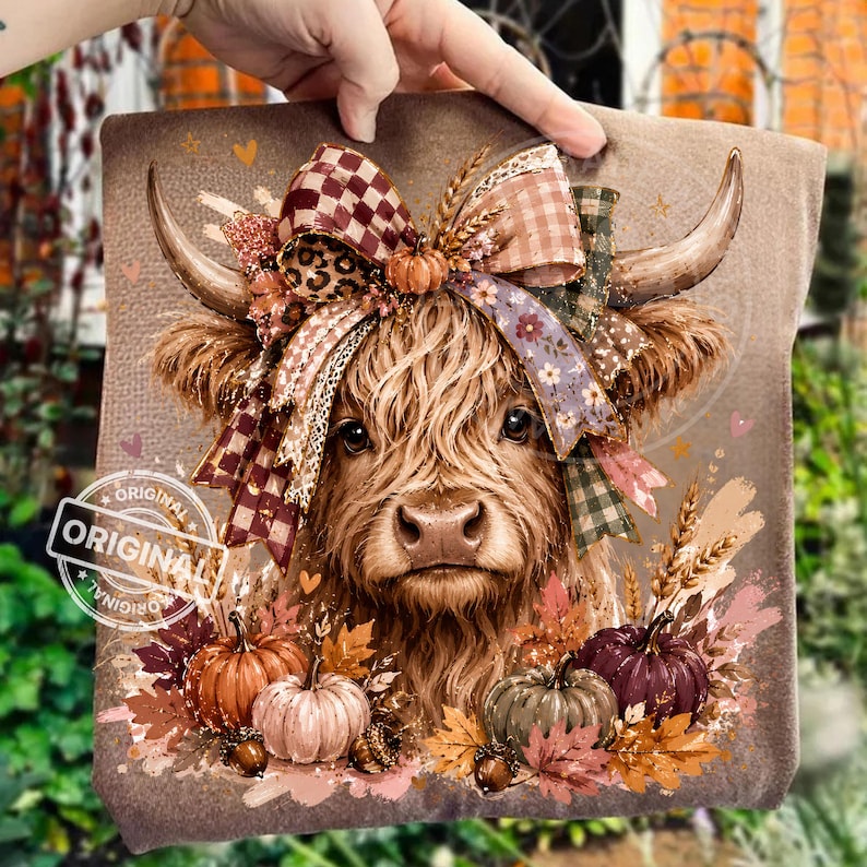 May include: A brown t-shirt featuring a graphic of a Highland cow with a large, multi-patterned bow between its horns. The cow is surrounded by autumn leaves, wheat stalks, and pumpkins in shades of orange, green, and purple. The design includes small hearts.