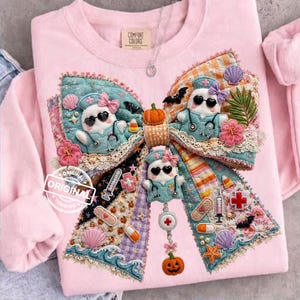 May include: Pink Comfort Colors sweatshirt with a large, decorative bow applique. The bow features three cartoon nurse figures, pumpkins, bats, and medical-themed embellishments. The word "ORIGINAL" is stamped on the sweatshirt.