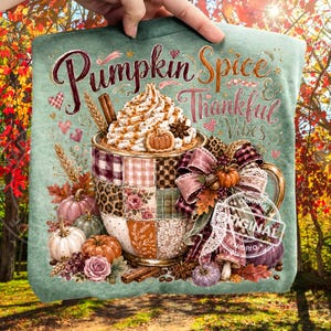Könnte beinhalten: Ein salbeigrünes T-Shirt mit einer Grafik einer Tasse, gefüllt mit Schlagsahne und einer Zimtstange. Der Text lautet Pumpkin Spice & Thankful Vibes. Die Tasse hat ein Patchwork-Design mit Kürbissen, Weizen und einer großen karierten Schleife.