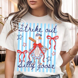 May include: White t-shirt with a baseball-themed graphic. The design features a goose wearing a baseball cap and holding a bat, with the words "Strike out" and "Silly goose" in blue script. Red bows and stars complete the design.