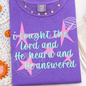May include: A purple Comfort Colors t-shirt with the text "i sought the lord and He heard and He answered" in light blue script. Pink starbursts accent the text. A gold butterfly necklace rests on the shirt.