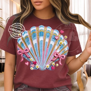 以下が含まれることがあります： 貝殻のデザインが施された、バーガンディ色のTシャツ。貝殻は青、ピンク、金で、ピンクのリボン、小さなハート、花が描かれています。「ORIGINAL」の文字がシャツの左上にプリントされています。