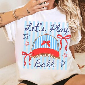 May include: White t-shirt with a graphic design featuring a baseball cap with blue and white stripes, a red brim, and a red bow. The text "Let's Play Ball" is written in blue script, with star and bow accents.