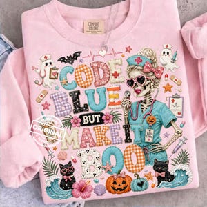 May include: Pink sweatshirt with a colorful graphic design. The design features the text "CODE BLUE BUT MAKE IT BOO" with a skeleton nurse, pumpkins, cats, and other Halloween-themed elements. The sweatshirt has a crew neck and long sleeves.
