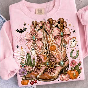 Puede incluir: Sudadera rosa con un diseño decorativo de botas de vaquero, lazos y joyas. El diseño incluye calabazas, fantasmas, una herradura y la palabra "ORIGINAL".