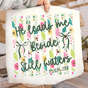 May include: A white throw blanket with a floral design and the text "He leads me beside still waters" in green script. The design includes pink, yellow, and green flowers and leaves, with vertical striped accents. Psalm 23:2 is also printed.