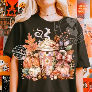 Könnte beinhalten: Ein schwarzes T-Shirt mit einer Grafik eines Kaffeebechers mit Schlagsahne, umgeben von herbstlichen Elementen wie orangefarbenen und weißen Kürbissen, Herbstblättern, Zimtstangen, Pilzen, Eicheln und einer großen rosa Schleife.