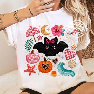 May include: A white t-shirt featuring a central graphic of a black bat with a leopard print bow, surrounded by various icons including a pink checkered umbrella, a crescent moon, a pink hibiscus, a ghost, a pumpkin, a wave, and a starfish.