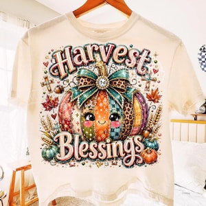 Op de afbeelding: Een crèmekleurig t-shirt met een afbeelding van een cartoonpompoen met een gezicht, een blauwgroene strik en luipaardprint accenten. De tekst Harvest Blessings staat in een decoratief lettertype rondom de pompoen en herfstbladeren gedrukt.