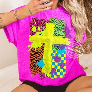 May include: A bright pink t-shirt featuring a neon yellow cross design. The cross is overlaid on a patchwork of animal prints and checkered patterns in vibrant colors like orange, green, and purple. The word "ORIGINAL" is stamped on the shirt.