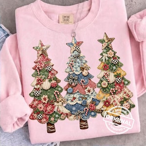 Puede incluir: Una sudadera de cuello redondo rosa claro con un gráfico de tres árboles de Navidad estilizados. Cada árbol está decorado con varios parches de tela estampados, incluyendo diseños florales, de cuadros y de lunares, rematados con una estrella.