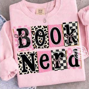 May include: A light pink sweatshirt with the words "BOOK NERD" in a playful design. The letters are black with various patterns, including leopard print and checkered, and accented with pink bows and gold glitter.