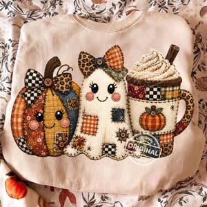May include: A light beige sweatshirt featuring a graphic print of a patchwork pumpkin with a bow, a patchwork ghost with a bow, and a mug of whipped cream with a cinnamon stick. The design uses various patterns like plaid, leopard, and floral.
