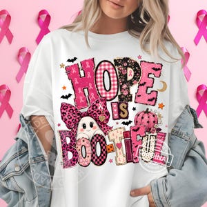 Op de afbeelding: Een wit t-shirt met een grafisch ontwerp met de tekst "HOPE IS BOO-TIFUL". Het ontwerp bevat een geest met een roze strik met luipaardprint, een roze pompoen, vleermuizen, sterren en roze lintpatronen in diverse texturen en ruitjes.