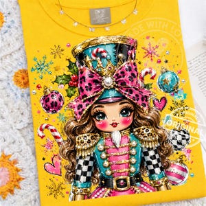 Può includere: Una t-shirt gialla con un'illustrazione grafica di un personaggio schiaccianoci dai capelli castani, che indossa un cappello e un'uniforme con stampa leopardata color verde acqua e rosa, circondato da decorazioni natalizie, fiocchi di neve e agrifoglio.