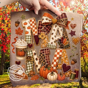 May include: A grey fabric surface featuring the word MAMA in large, patterned letters. The design includes a coffee cup, pumpkins, acorns, leaves, hearts, stars, and a bow. The letters are decorated with plaid, leopard print, and floral patterns.