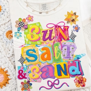 Peut inclure: T-shirt crème avec l'inscription "SUN SALT & SAND" en lettres colorées de style patchwork. Le motif comprend des accents floraux et géométriques, un nœud rose et un collier délicat.