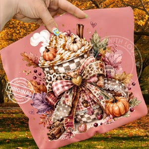 May include: A pink fabric square featuring a graphic illustration of a coffee cup topped with whipped cream, cinnamon sticks, and small pumpkins. The cup is decorated with a large, multi-patterned bow, autumn leaves, and berries.