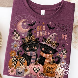 May include: Purple t-shirt featuring two black cats wearing witch hats and scarves, surrounded by pumpkins, flowers, and a crescent moon. The shirt has the text "Boo Babe" and "Stay Spooky".