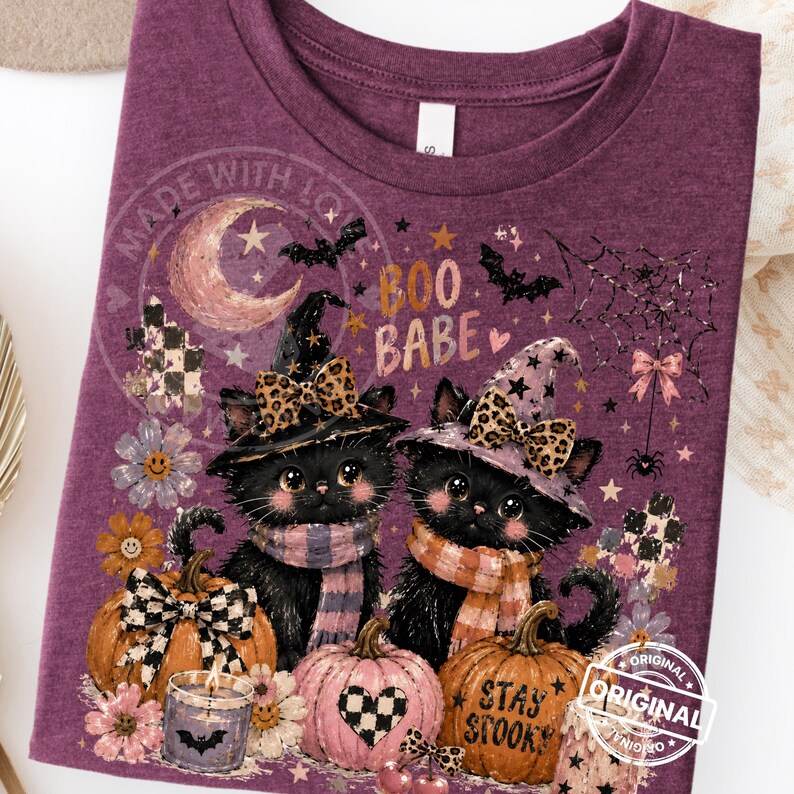 May include: Purple t-shirt featuring two black cats wearing witch hats and scarves, surrounded by pumpkins, flowers, and a crescent moon. The shirt has the text "Boo Babe" and "Stay Spooky".