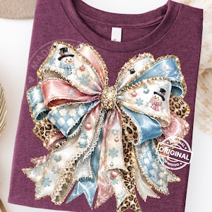 May include: A maroon t-shirt featuring a large, central graphic of a multi-layered bow. The bow design incorporates light blue, pink, and leopard print patterns, accented with illustrations of snowmen, snowflakes, and ornaments. The word ORIGINAL is printed in a circular emblem on the lower right.