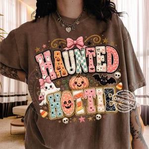 May include: A brown t-shirt featuring a graphic print with the text HAUNTED HOTTIE. The design includes a ghost with a pink bow, a jack-o'-lantern, a bat, skulls, candy corn, and stars, all rendered in a quilted, stitched-style pattern.