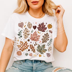 Può includere: Una t-shirt bianca a maniche corte con una stampa grafica centrale di varie foglie autunnali in diversi motivi, tra cui stampa leopardata, scozzese e toni della terra, intervallate da piccole ghiande e piccoli accenti a stella dorati.