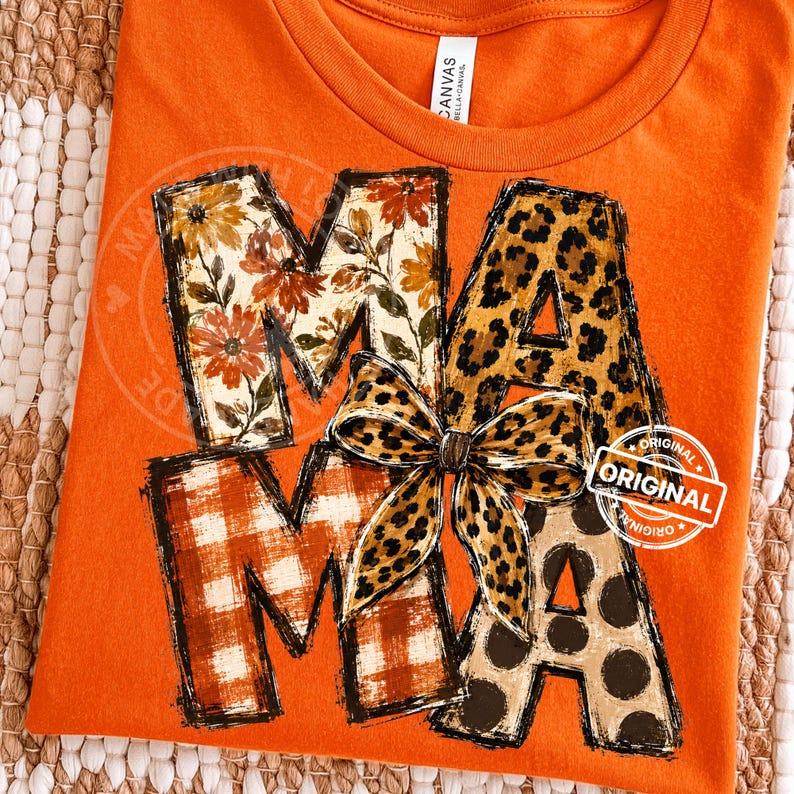 Puede incluir: Una camiseta naranja con la palabra MAMA en letras grandes. La M tiene un estampado floral, la A un estampado de leopardo, la segunda M un estampado de cuadros rojos y blancos, y la &uacute;ltima A un estampado de lunares marrones.