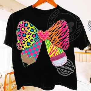 Op de afbeelding: Zwart T-shirt met een groot, kleurrijk strikdesign. De strik heeft verschillende patronen, waaronder luipaardprint, zebra strepen en een geruit patroon, met een hartvormig middenstuk. Het ontwerp is op het shirt gestikt.