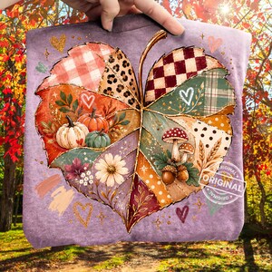 May include: A light purple t-shirt featuring a large, central leaf graphic composed of various patterned segments including plaid, leopard print, and polka dots. The leaf design incorporates illustrations of pumpkins, mushrooms, acorns, and flowers.