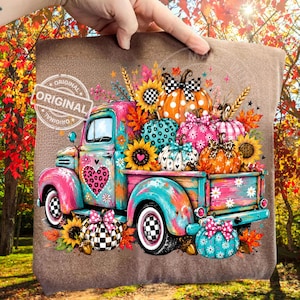 May include: A brown t-shirt featuring a colorful graphic of a vintage pickup truck filled with patterned pumpkins. The truck is teal and pink with a leopard print heart. The pumpkins feature various designs including polka dots, plaid, and leopard print.