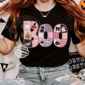 May include: A black short-sleeved t-shirt featuring the word BOO in large, patchwork-style letters. The letters are filled with pink, white, and black patterns including ghosts, pumpkins, bats, skulls, spiders, and bows in a quilted design.