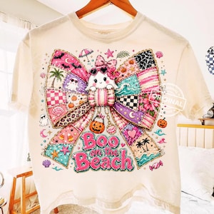 May include: Cream-colored t-shirt featuring a colorful bow design with a ghost wearing sunglasses. The bow is made of patchwork patterns with beach and Halloween elements. The text "Boo on the Beach" is displayed in pink.