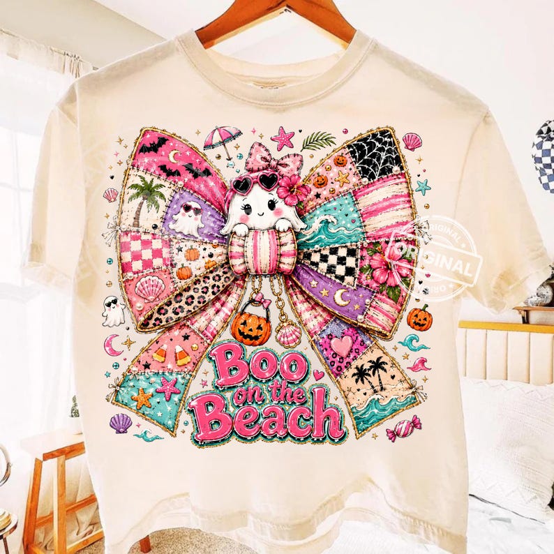 May include: Cream-colored t-shirt featuring a colorful bow design with a ghost wearing sunglasses. The bow is made of patchwork patterns with beach and Halloween elements. The text "Boo on the Beach" is displayed in pink.