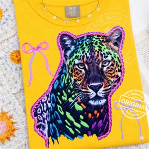 Könnte beinhalten: Ein gelbes T-Shirt mit einem farbenfrohen Leopardenmuster, das pink umrandet ist. Die Flecken des Leoparden sind eine Mischung aus Grün, Blau, Orange und Lila. Das Shirt hat ein Comfort Colors-Etikett und eine rosa Schleife. Der Text "ORIGINAL" ist ebenfalls sichtbar.