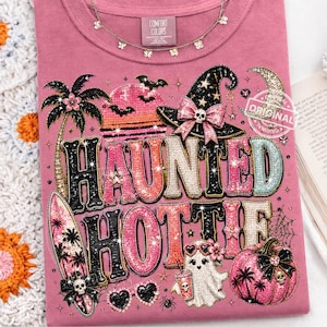 以下が含まれることがあります： ピンク色のTシャツ。スパンコールで「HAUNTED HOTTIE」の文字が描かれています。魔女の帽子、お化け、カボチャ、サーフボードなど、ハロウィーンをテーマにしたグラフィックが特徴です。上部にはシルバーの蝶のネックレスがあります。