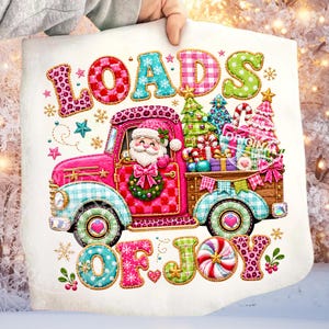 May include: A white fabric square featuring a colourful graphic of a pink truck driven by Santa Claus. The truck carries Christmas trees and gifts. The text reads "LOADS OF JOY" in patterned letters. The design includes snowflakes and stars.