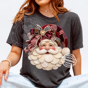 May include: A dark heather gray t-shirt featuring a graphic of Santa Claus wearing a patchwork-style hat with a large bow. The hat includes red, burgundy, and black-and-white checkered patterns. Santa has a white beard and a gold heart charm.