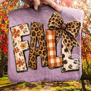 May include: A heathered purple t-shirt featuring the word FALL in large block letters. The F has an orange floral pattern, the A has a leopard print, the L has an orange and white buffalo plaid, and the second L has brown polka dots with a large leopard print bow attached.