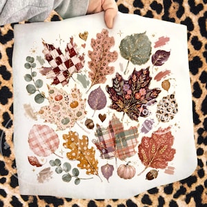 May include: A white square transfer sheet featuring a collection of autumn-themed illustrations including various leaves with plaid, leopard print, and floral patterns, small pumpkins, acorns, and eucalyptus sprigs, accented with gold sparkles.