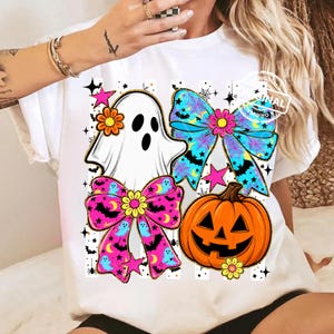 以下が含まれることがあります： ハロウィーンのデザインが施された白いTシャツ。デザインには、お化け、2つのリボン、ジャック・オー・ランタン、星が含まれています。リボンはピンクと青で、月とコウモリの模様です。お化けは頭に花を持っています。