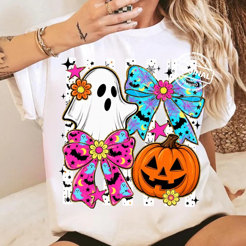 以下が含まれることがあります： ハロウィーンのデザインが施された白いTシャツ。デザインには、お化け、2つのリボン、ジャック・オー・ランタン、星が含まれています。リボンはピンクと青で、月とコウモリの模様です。お化けは頭に花を持っています。