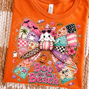 May include: Orange t-shirt with a colorful graphic featuring a ghost wearing sunglasses, a patchwork bow, and the text "Boo on the Beach." The design includes Halloween and beach-themed elements like pumpkins, seashells, and palm trees.