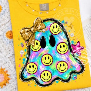 May include: A bright yellow t-shirt featuring a central graphic of a ghost shape filled with a multicoloured tie-dye pattern and several yellow smiley faces. The ghost has a large gold glitter bow on its head and a pink star on its right side.