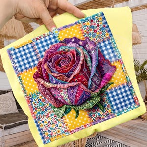 Könnte beinhalten: Ein dekoratives Kissen mit einer lebendigen, mehrfarbigen Rosenapplikation. Die Rose besteht aus verschiedenen gemusterten Stoffen, darunter Blumen-, Tupfen- und Vichy-Muster. Das Kissen hat einen gelben Rand und ein Patchwork-Design, das einen einzigartigen und künstlerischen Look erzeugt.