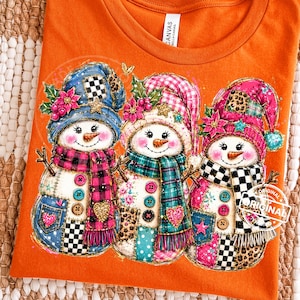 Puede incluir: Una camiseta naranja con un estampado gráfico de tres muñecos de nieve que llevan gorros y bufandas coloridos con varios patrones, incluyendo cuadros, estampado de leopardo y ajedrez. Están decorados con botones, corazones y flores de pascua.