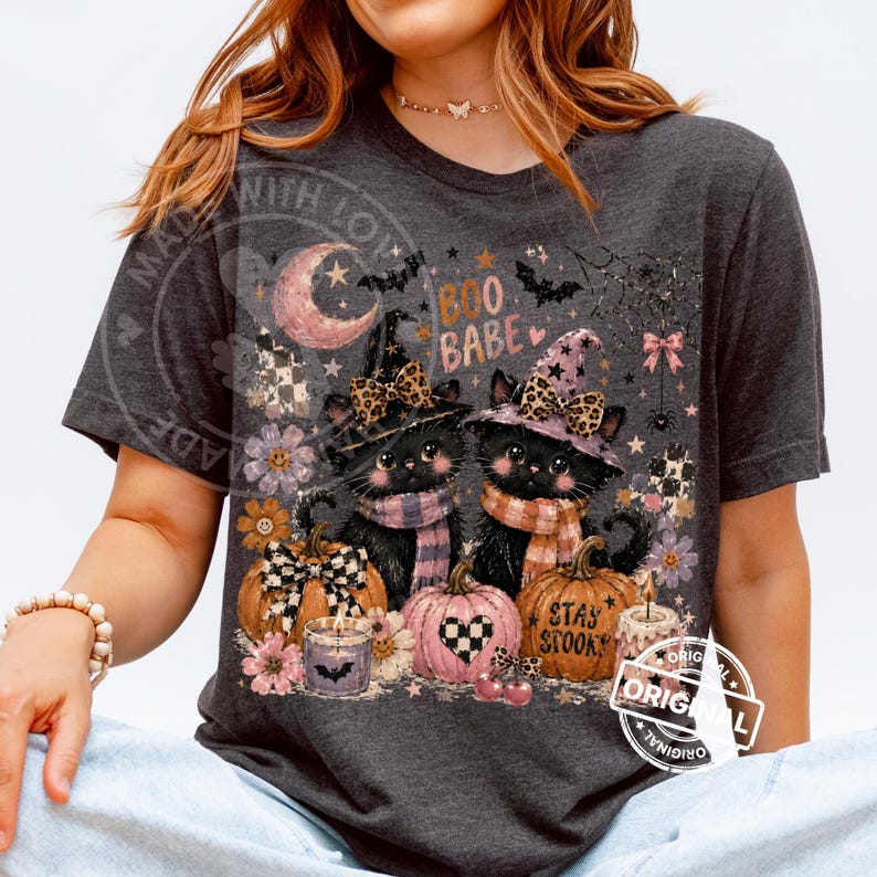 May include: Dark gray t-shirt featuring a Halloween-themed graphic. The design includes two black cats wearing witch hats and scarves, surrounded by pumpkins, candles, and the text "Boo Babe" and "Stay Spooky".
