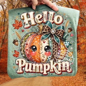 Puede incluir: Una camiseta verde salvia con un gráfico de una calabaza de dibujos animados con ojos grandes, mejillas sonrosadas y un lazo con estampado de leopardo. El texto "Hello Pumpkin" está impreso en una fuente decorativa. Incluye hojas de otoño, trigo y bellotas.