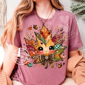 Peut inclure: Un t-shirt mauve à manches courtes orné d'un grand motif central représentant une feuille d'érable de dessin animé avec un visage souriant, de longs cils et un nœud marron foncé. La feuille présente des motifs léopard, carreaux, citrouilles.